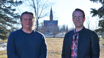Två män står framför en gräsmatta. Solen skiner och i bakgrunden synns Sibbo kyrka.