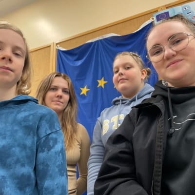 Topias Huhtiniemi, Sara Jauhiainen, Iita Karppinen ja Siiri Kantonen katsovat alaviistosti kameraan ja hymyilevät. Taustalla on luokkahuoneen seinää ja EU-lippu.
