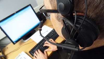 En person vid med hörlurar och mikrofon sitter vid ett arbetsbord framför en dator.