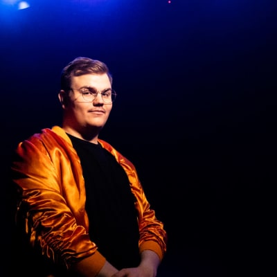 UMK-finalisti Aksel Kankaanranta seisoo tummansinisen taustan edessä kädet sylissä. Silmälasipäisellä Kankaanrannalla on yllään oranssi satiininen takki ja musta paita alla. 