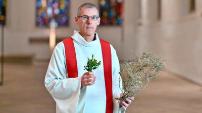 En man i en vit kåpa med röd skarf. Han har gröna kvistar i handen och står i en kyrka.