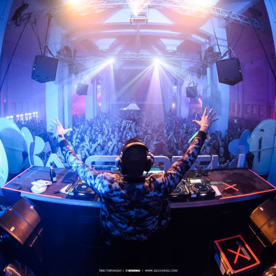 DJ seisoo soittopöydän takana kädet ilmassa, edessä näkyy kädet ilmassa juhliva täysi yleisö.