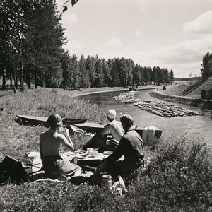 Kanavapiknikillä joskus 1950-luvulla. Mustavalkoinen kuva: Kolme henkilöä piknikillä kanavan rannalla, kanavassa uitetaan tukkeja.