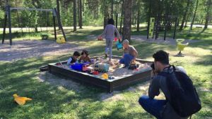 Barn lekte på gården i Mjölbolsta sommaren 2016. Fotografen Ahmed Alalousi bodde också på förläggningen. Han har nu uppehållstillstånd i Finland. Barn leker i en sandlåda. En ung man med ryggsäck på ryggen fotograferar dem. Sommar, solen skiner. I bakgrunden syns gungor och klätterställningar.