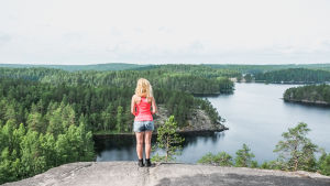 Malin i Repovesi, ett av favoritresmålen i Finland. En kvinna iklädd shorts och ett rosa linne står på en bergskant och blickar ut över en sjö och skog i Finland. Det ser mycket fridfullt ut.