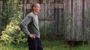 Ett självporträtt från 1982. Veijo Rönkkönen står iklädd joggingbyxor och tröja i högt gräs. I bakgrunden en grånad lada med mossigt tak.