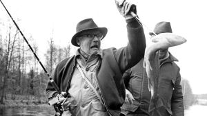 Presideanta Urho Kekkonen lei guollebivdi. Urho Kekkonen painii kalan kanssa hämmästynyt ilme kasvoillaan.