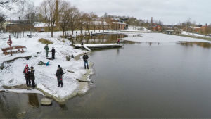 Helsingin Vanhankaupunginlahti on luontohelmi, josta kaupunkilaiset saavat virkistystä. Ihmisiä kalastamassa rannalla. Maassa on hento lumipeite, vesi on sulaa. Taustalla on taloja.