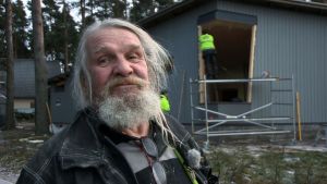 Dale myhäilee tyytyväisenä, ja taustalla lasiasentajat valmistautuvat suureen urakkaan. Mies lähikuvassa rakennuksen edessä