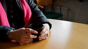 En kvinna sitter vid ett bord med en kaffekopp i händerna. Kvinnans ansikte syns inte på bilden.
