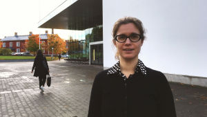 Katarina Hellen forskar i hur man uppnår långsiktig lycka. Katarina Hellen står utomhus i höstsolen med universitetsbiblioteket tritonia bakom sig. Hon bär en svart tröja och har svartbågade glasögon.