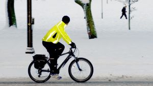 Det finns inget dåligt cykelväder. En man iklädd en neongul cykeljacka, cyklar på en snötäckt väg. I bakgrunden syns en snöklädd park och en man som går.