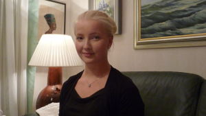 Michelle Gustafsson sitter i en soffa i sitt hem. Tavlor och bordslampa i bakgrunden. Kväll