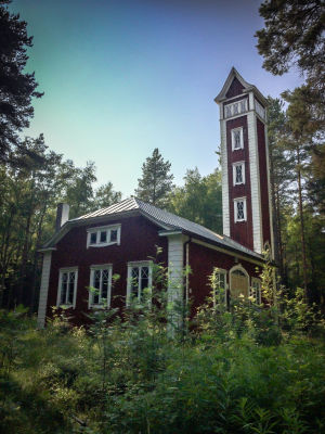 I stället för brandmän är det en spillkråka som huserar här. Ingen siren ljuder längre, bara ett rytmiskt knackande. Ulkokuva paloasemasta, vanha punamullalla maalattu rakennus, valkoiset nurkkalaudat ja ikkunanpielet, ympärillä kesäistä metsää, kuvattu Varjakansaaren vanhassa sahakylässä.