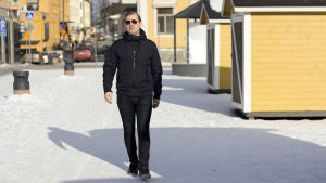 En dag i taget, då kommer man vidare. En man som går på en trottoar. Det är en solig vinterdag och han bär solglasögon. I bakgrunden syns flera gula hus.