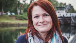 Reetta Nousiainen on erittäin viehättynyt kotikylästään ja on paneutunut sen historiaan innolla. Punahiuksinen nainen jolla tummansininen villatakki ja kukallinen huivi kaulassa hymyilee kohti kameraa. Taustalla vettä.