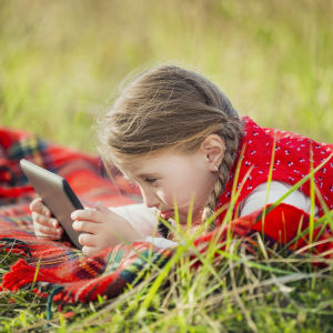 Tyttö makaa nurmikolla tablettitietokoneen kanssa.
