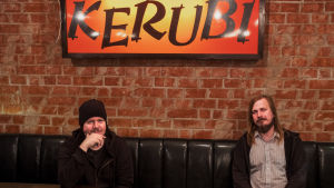 Musikerna Aki Tykki och Ville Karttunen sitter i svart lädersoffa, ovanför dem neonskylt med texten Ravintola Kerubi.