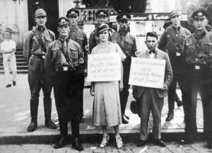 Saksalaisnainen ja juutalaismies julkisesti nöyryytettävinä, ympärillä SA- ja SS-miehiä SA- ja SS-miehet nöyryyttävät julkisesti juutalaismiestä ja saksalaisnaista, jotka on tavattu yhdessä