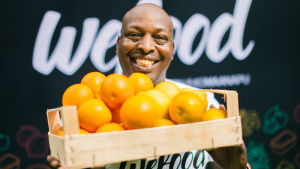 Wefood planerar att öppna en matsvinnsbutik i Helsingforsregionen i år. Den före detta löparen Wilson Kirwa fungerar som ett av projektets ansikten utåt. Före detta löparen Wilson Kirwa håller i en trälåda med apelsiner i. I bakgrunder står det Wefood.