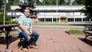 Jan-Erik Till jobbar som lärare i Karis. Man sitter på en röd bänk framför en rappad skolbyggnad.