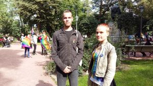 Jim Töyrylä och Linn Gröndahl ung man i svarta kläder och ung kvinna i ljusa kläder står framför Porthans staty i Åbo. I bakgrunden människor med regnbågsflaggor