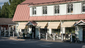 Kylän ravintola ilta-auringossa. Vanha valkoinen puutalo jolla punainen peltikatto, talossa ravintola ja talon edustalla terassi.