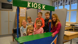 En del av skolans elever samlade i den egenhändigt tillverkade kiosken. Glada elever bakom i en leksakskiosk.