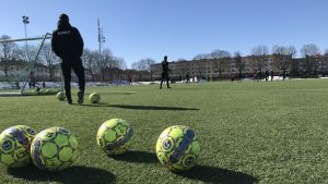 Fotbollsträning Konstgräsplan utomhus med fotbollar i förgrunden och en man med texten "Dalkurd FF" på ryggen.