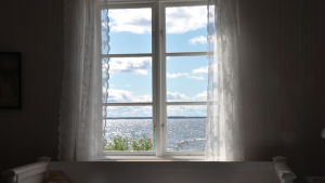 Havsutsikt från köksfönstret är det bästa, men den kostar. Havsutsikt från ett fönster. Vita tyllgardiner, sommar, glittrande hav.