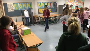 Efter att eleverna röstat stämplas röstsedeln och de får lägga den i valurnan. Elever står i ett klassrum som är en vallokal.