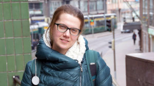 Maria Eliseeva iklädd mörkgrön täckjacka står och tittar rakt in i kameran med några stora våningshus i bakgrunden.