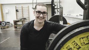 Elin Hassel - din tunga vikt-peppare. En kvinna sitter bredvid en skivstång med en vikt på. Hon ler.