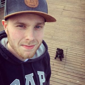 Nicos trogne vän som alltid ställer upp. En man i keps står på en terass och tittar in i kameran. I bakgrunden sitter en liten svart hund av rasen mops.