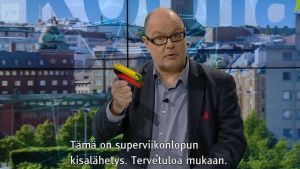 Pressiklubi ohjelmatekstitetään ja sitä myös tulkattiin viittomakielelle. Kuvassa juontaja Ruben Stiller. Ruben Stiller katsoo kohti ja pitää kädessä kisayleisön käyttämää räikkää
