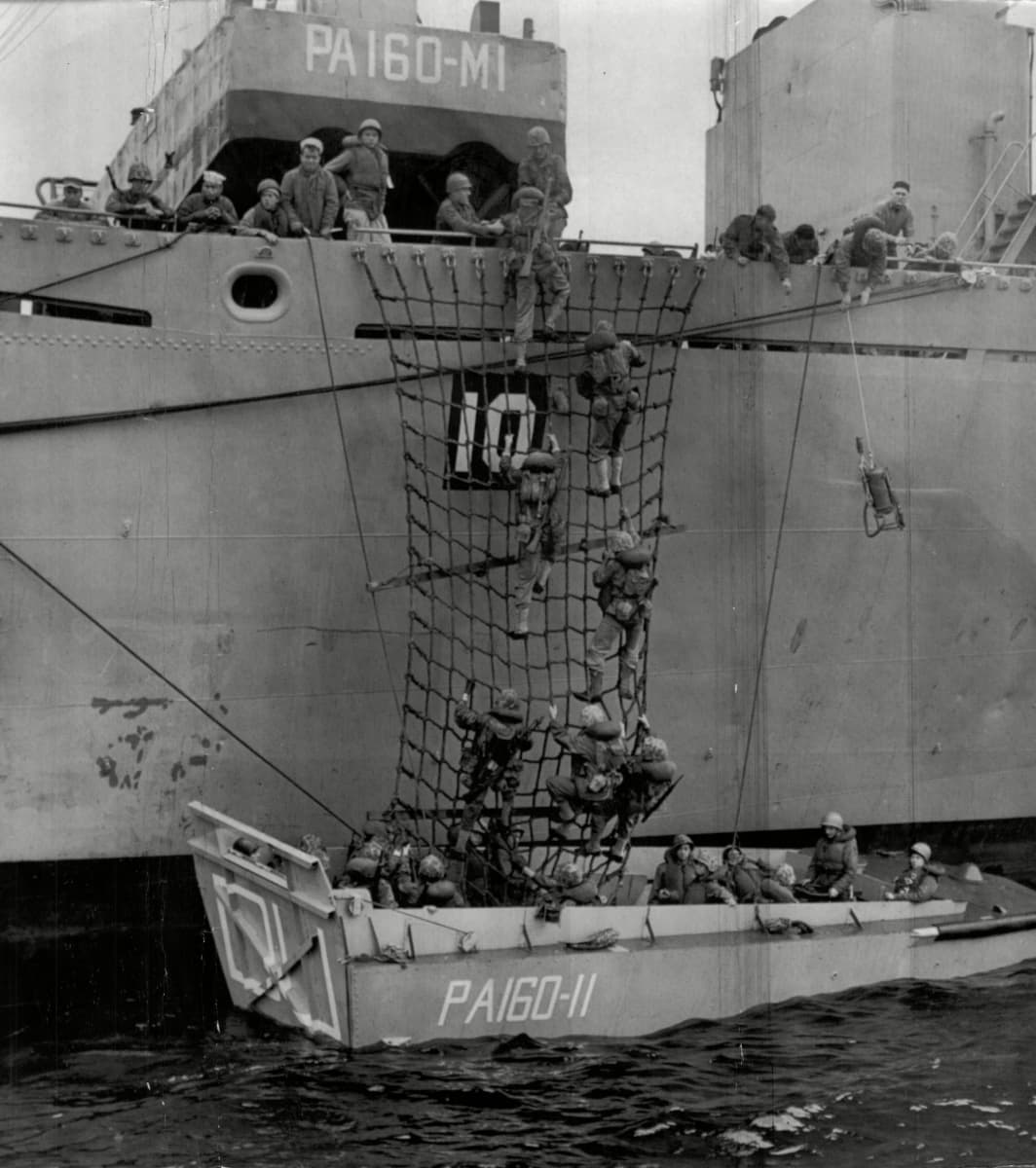 Mustavalkokuvassa amerikkalaisia sotilaita kypärät päässä laskeutuu verkkoa pitkin sota-aluksesta maihinnousuveneeseen.