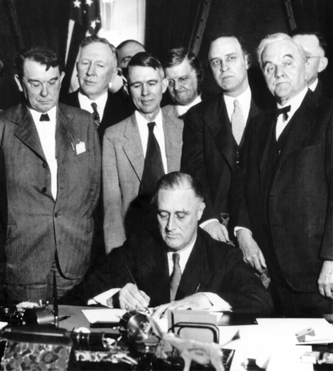 Mustavalkoisessa kuvassa presidentti Franklin Delano Roosevelt allekirjoittaa paperin työpöytänsä ääressä. Taustalla on miehiä puvuissa seuraamassa allekirjoitusta.
