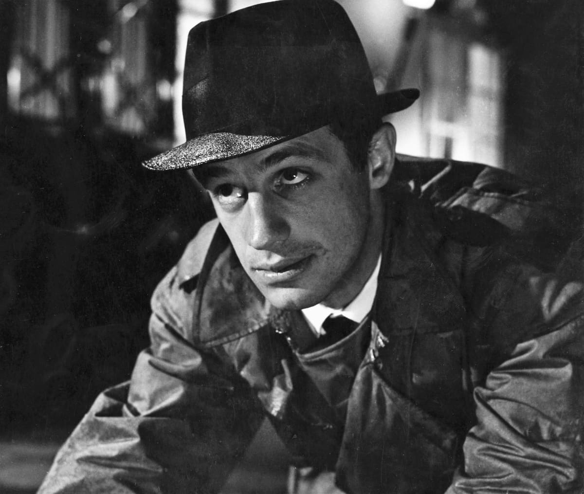 Tummassa mustavalkokuvassa Belmondolla on päässään sateen kastelema hattu ja trench-coat. Hän katsoo hieman yläviistoon.