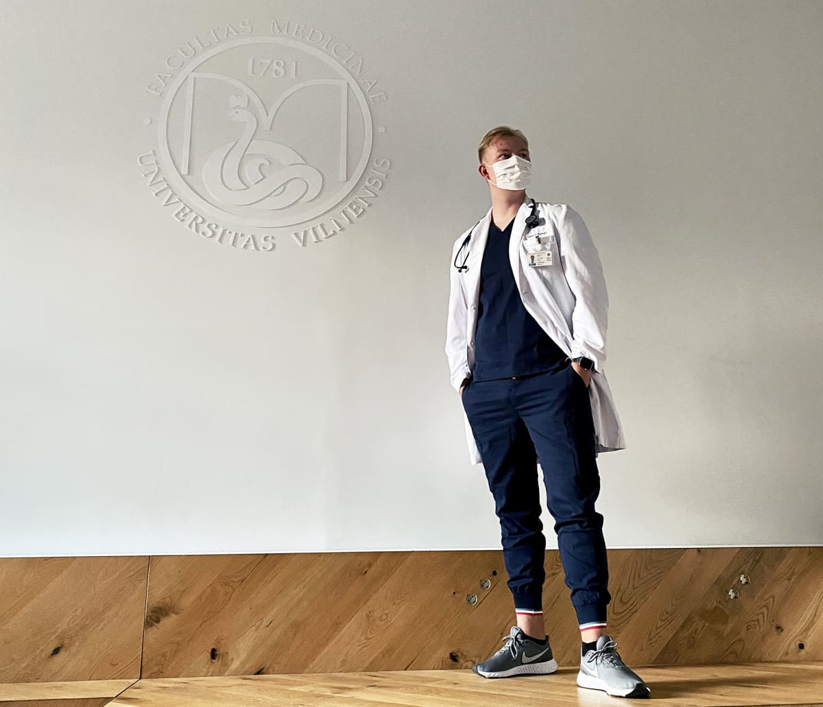 Nuori mies lääkärinvaatteissa seisoo Vilnan yliopiston logon alla.
