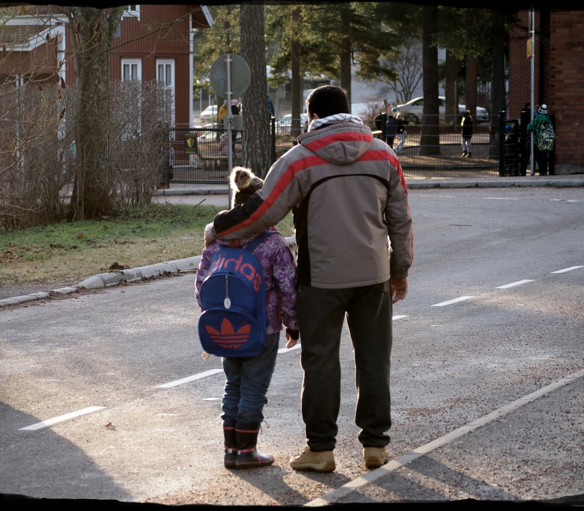 Far håller arm om dotter, de står med ryggen till och ser på avstånd på en skola
