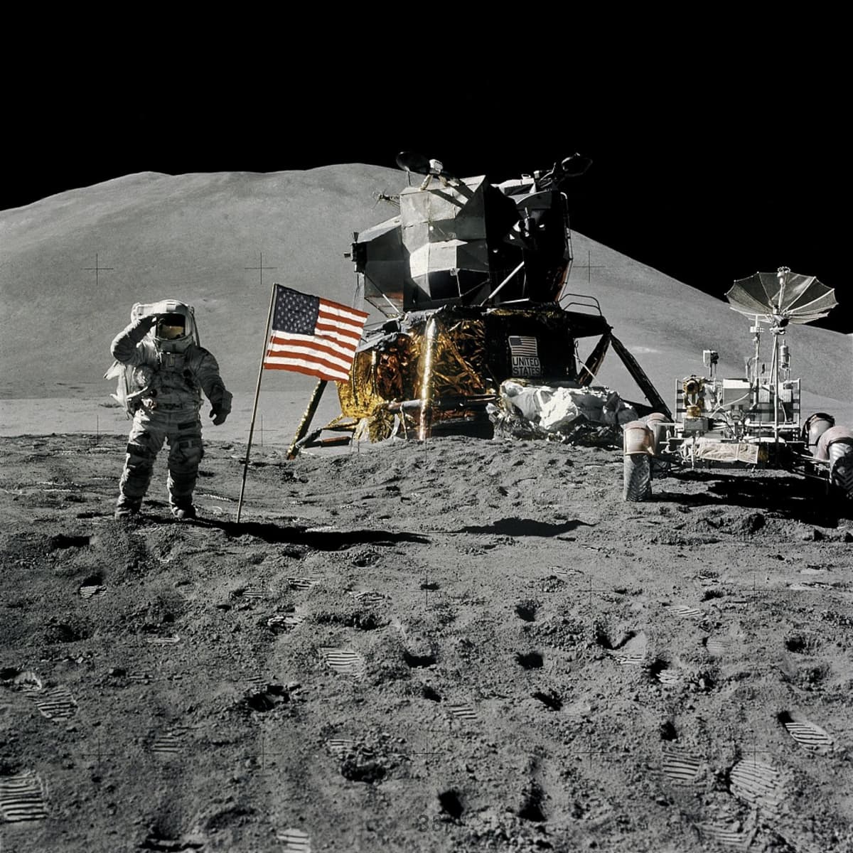 Astronautti Kuun pinnalla, edessä pajon jalanjälkiä, takana Yhdysvaltain lippu, laskeutuja ja mönkijä. Taustalla kukkula.