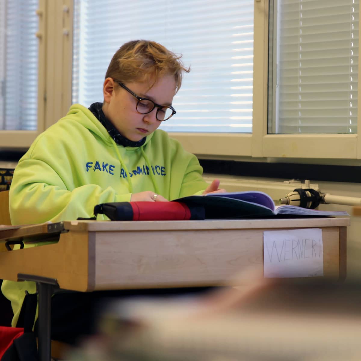 Silmälasipäinen Werneri Lappi istuu neonkeltainen huppari päällään pulpetissa ja tekee kirjasta tehtäviä. Pulpetin etuosassa on kyltti, jossa lukee Werneri.