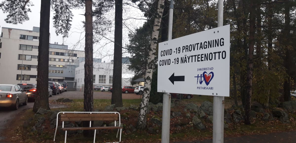 Covid -19 -näytteenottoon viittaava kyltti Pietarsaaren terveyskeskuksen pihamaalla, autojono on pitkä.