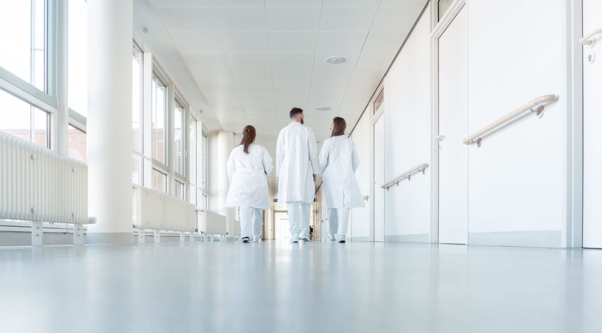Tre läkare klädda i vita rockar går i en sjukhuskorridor. De har ryggarna vända mot kameran.