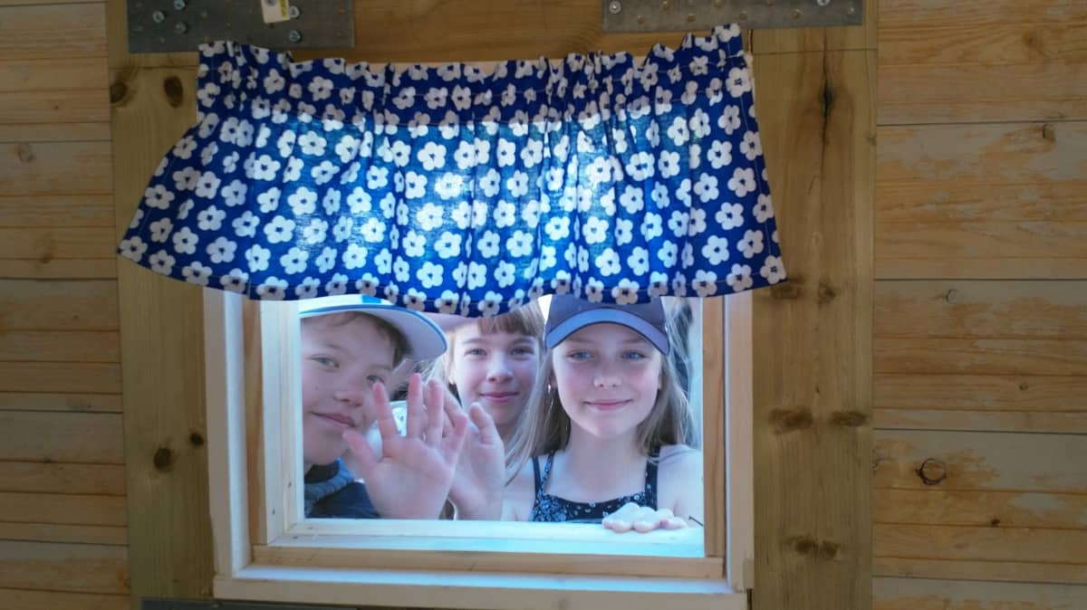 Kuvassa kolme lasta vilkuttaa leikkimökin ikkunasta