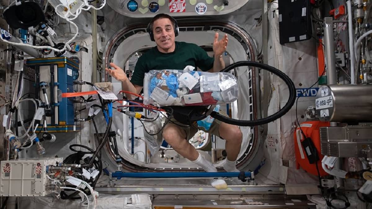 Shortseihin pukeutunut mies leijuu avaruusasemalla. Edessään hänellä on iso muovipussi täynnä roskia. 
