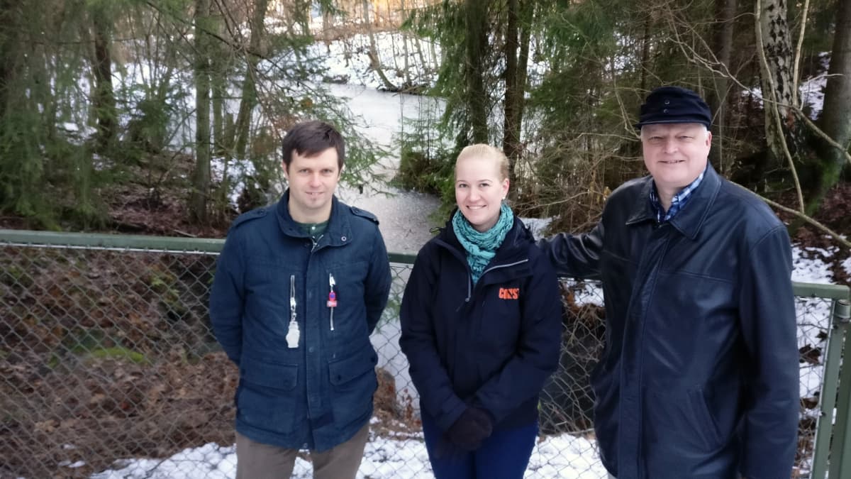 Metsittyneen kaivannon edessä seisovat arkeologi Andreas Koivisto sekä Linnaisten omakotiyhdistyksestä Minttu Lukkala ja Jukka Mäkinen.  Kuvassa näkyy myös kaivonnon ympäröivä verkkoaita. Vedellä täyttynyt kaivanto, joka on jäässä.