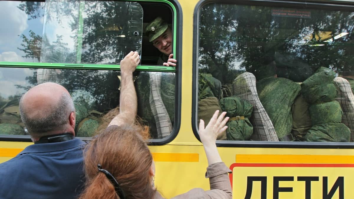 Varusmiehiä istuu keltaisessa bussissa, yksi heistä kurkistaa ikkunasta ulos. Vanhempi mies ja nainen vilkuttavat ulkona. Bussin kyljessä lukee venäjäksi: Deti eli Lapsia.