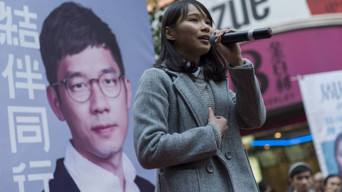 Harmaaseen päällystakkiin pukeutunut Agnes Chow puhuu mikrofoniin vaalijulisteen edessä.