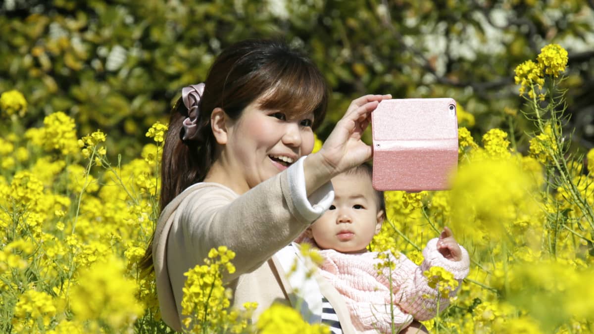 Nuori äiti ottaa puhelimella kuvaa itsestään ja vauvastaan kukkivien sinappikasvien keskellä.